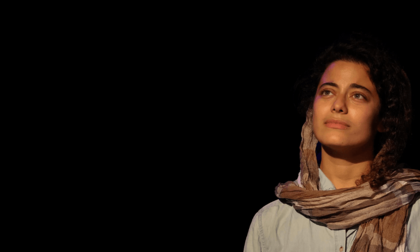 A woman with curly hair, wearing a light shirt and a scarf, looks thoughtfully upward. She is lit by a warm light against a dark, empty background, creating a dramatic and contemplative mood.