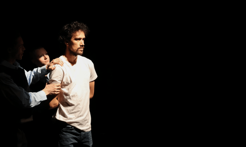 A man in a white t-shirt stands in a dark, spotlighted area, looking serious. Two people hold his arms from behind, partially hidden in the shadows. The scene is dramatic and tense.