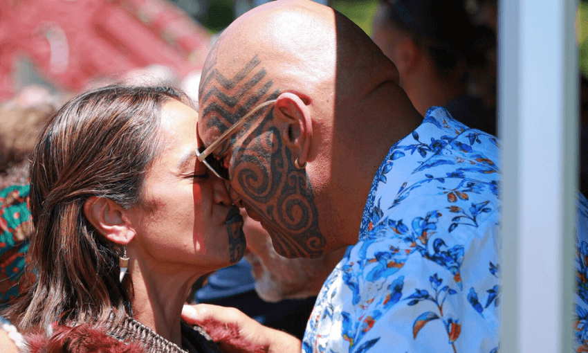 Two people with traditional Māori facial tattoos greet each other by pressing their noses and foreheads together, wearing patterned clothing, outdoors in bright sunlight.