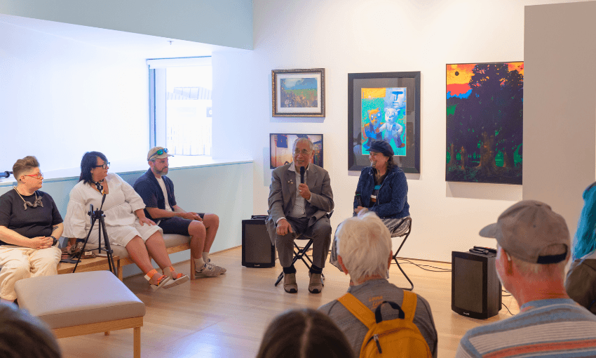 A group of people sit in a brightly lit art gallery, listening to two speakers with microphones. Colorful artwork hangs on the walls behind them. Some audience members wear hats and casual clothing.