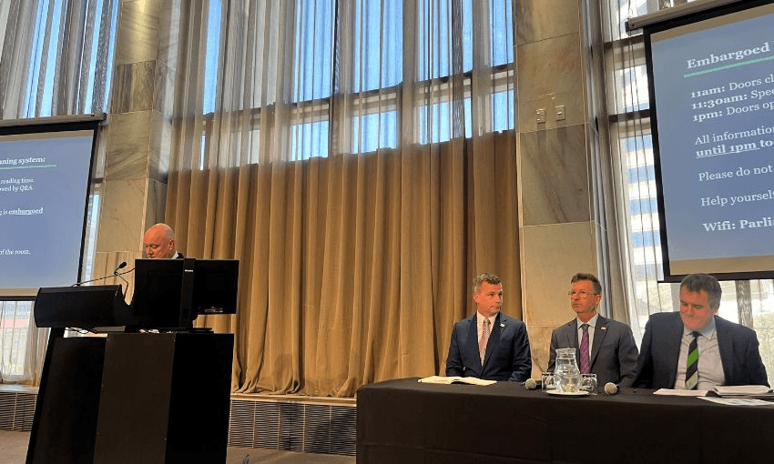Christopher Luxon's head is visible as he speaks at a podium. Beside him is a table seating David Seymour, Simon Court and Chris Bishop.