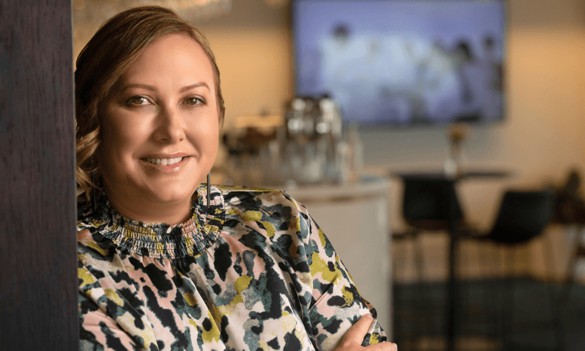 A woman with light brown hair and a patterned blouse smiles while leaning against a wooden surface in a modern, softly lit office or cafe setting. The background is blurred, showing tables and chairs.