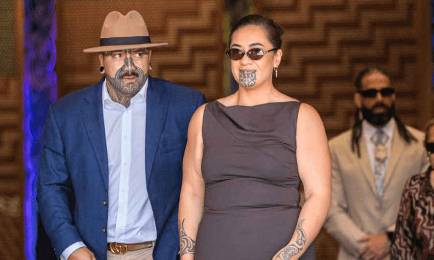 A man and a woman with Māori tattoos on their faces and arms walk together. The man wears a tan hat, blue suit, and white shirt. The woman wears sunglasses and a brown dress. Two people are blurred in the background.
