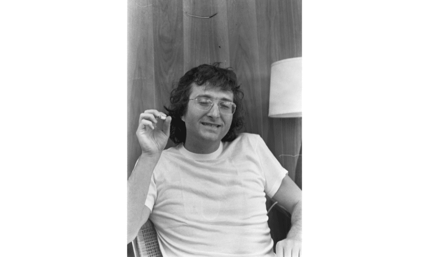 A black and white photo of a man with glasses, smiling, wearing a white t-shirt and holding a cigarette.