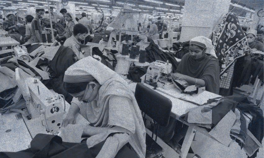 A group of women wearing headscarves work at sewing machines in a busy garment factory, surrounded by fabric and clothing pieces under bright overhead lights.
