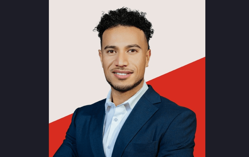 A man in a navy blue suit and white shirt smiles at the camera. He has short curly hair and light facial hair. The background is divided diagonally, with off-white on top and red on the bottom.