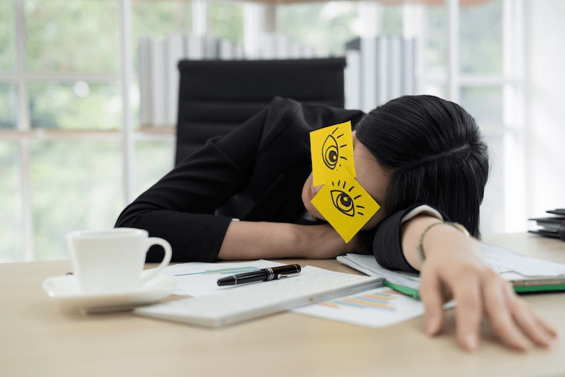 A person in business attire naps at a desk with their head resting on papers, wearing sticky notes with cartoon eyes drawn on them over their closed eyes. A coffee cup, pen, and keyboard are nearby.