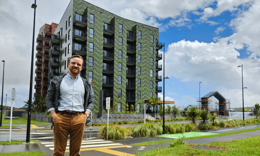 A smiling man stands on a sidewalk in front of a modern, multi-story apartment building on a bright, partly cloudy day. Lush greenery and a crosswalk are visible in the foreground.