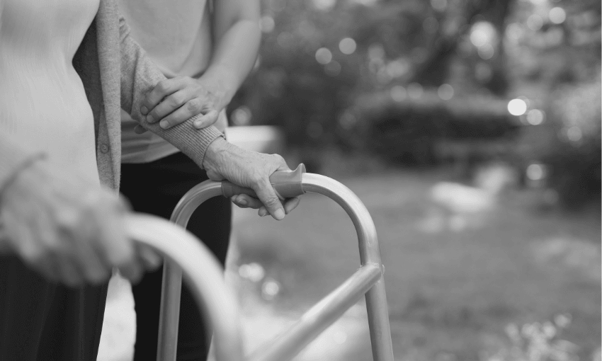 A close-up of an elderly person using a walker outdoors, with another person gently supporting them by holding their arm. The scene is in black and white.