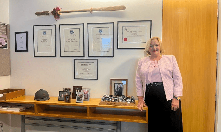 Judith Collins smiles while standing in her office, next to a wall of her framed degrees and gifted medals.