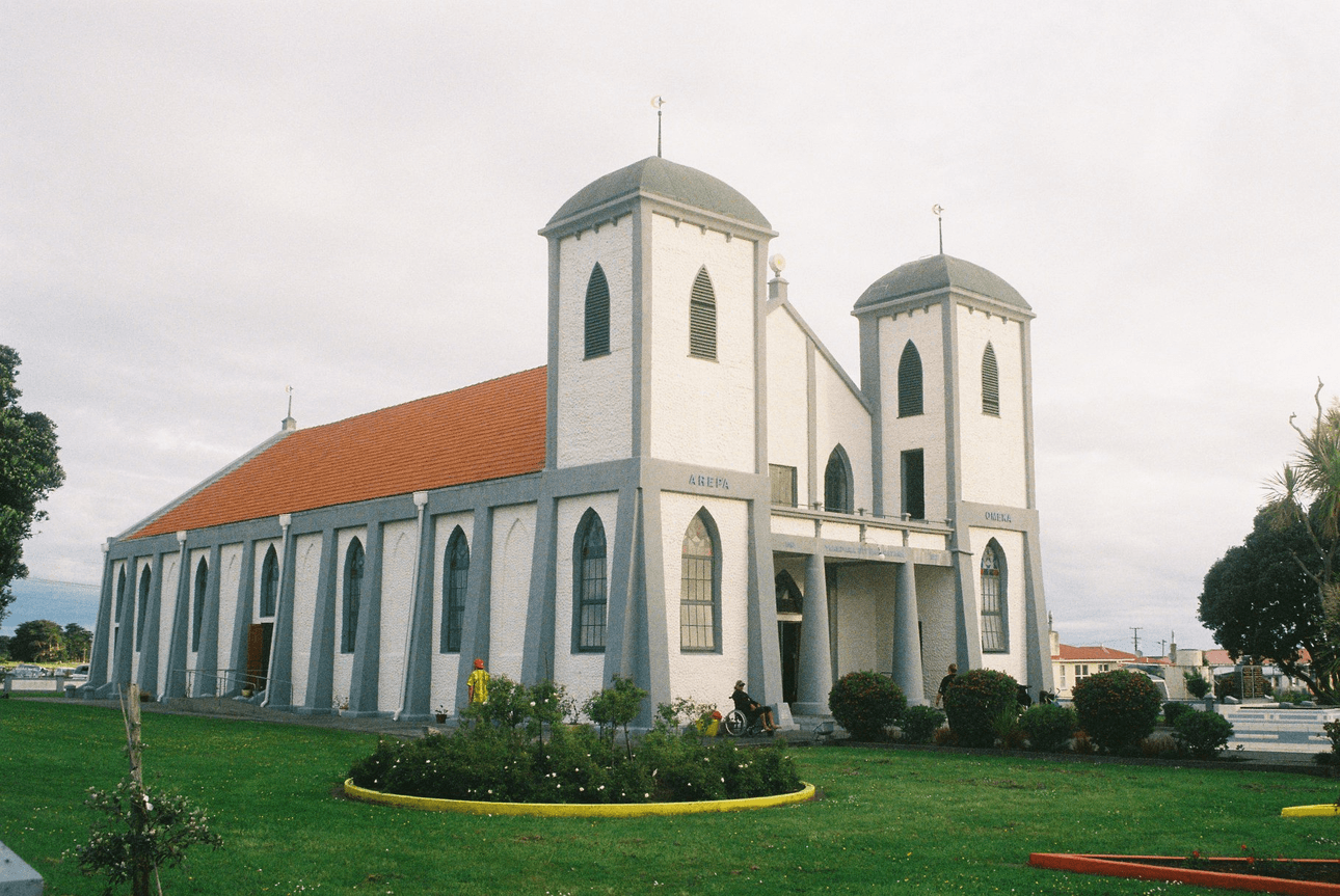 A white church with two tall towers, a red roof, arched windows, and green doors, sits on a well-kept lawn with a flowerbed in front. The sky is overcast and houses are visible in the background.