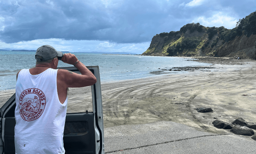 A man in a white tank top and gray cap stands by an open car door, looking out at a deserted, cloudy beach with tracks in the sand and green hills in the distance.