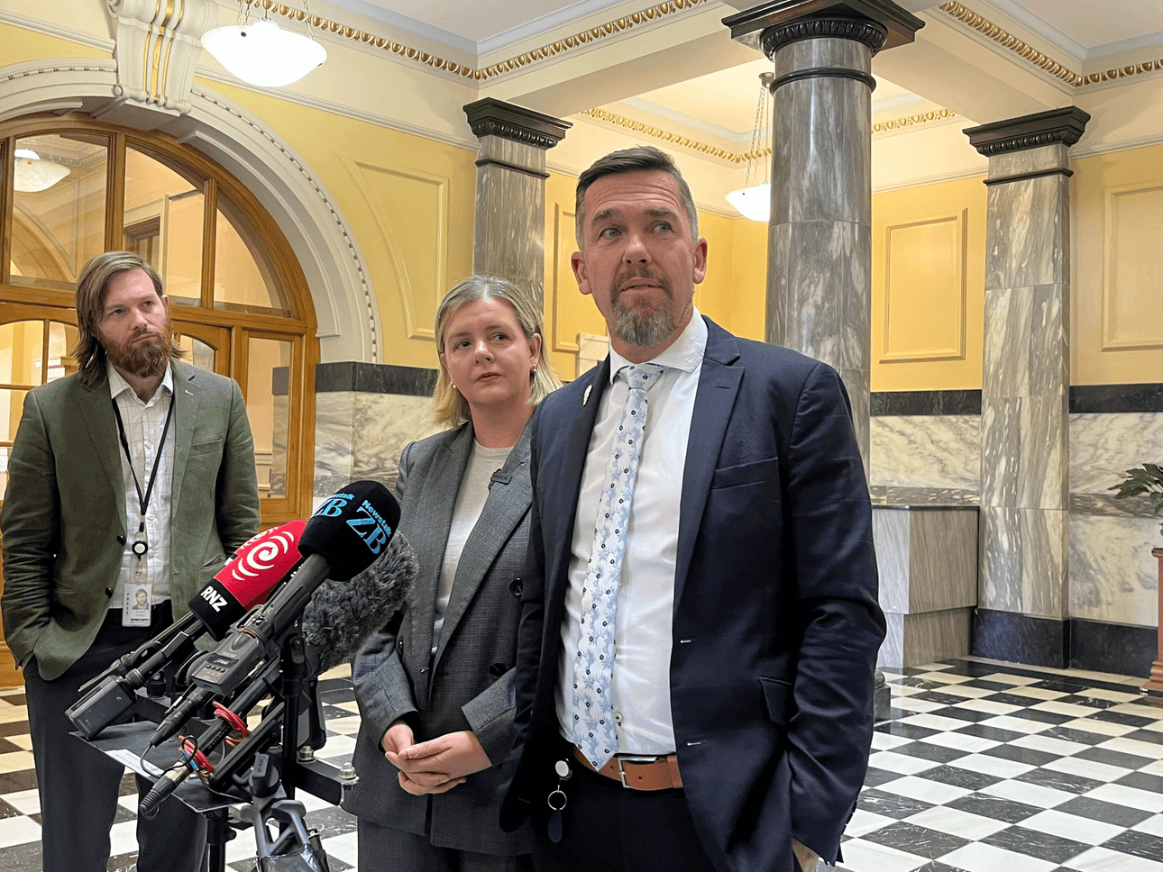 a tall man in a blue tie and a shorter blonde woman wearing a grey blazer look sideways with a bevy of microphones in front of them and distinctive black and white parliamment tiles behind them.