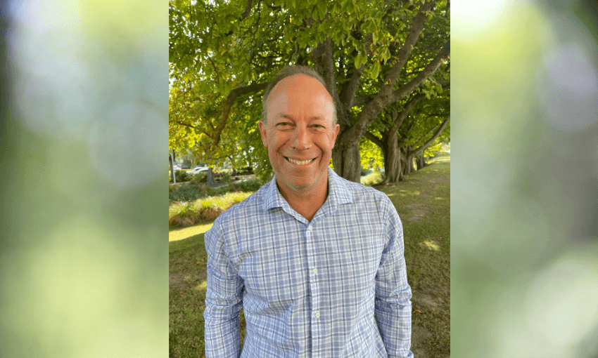 an older pakeha man in a light blue checked shirt smiling with his hands in his pockets and trees in the background