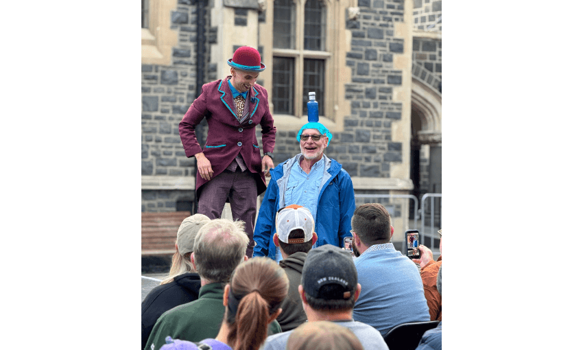 A photograph of an outdoor performance showing a circus performer balancing a bottle on the top of a man's head. A crowd looks on, some are filming with iPhones.
