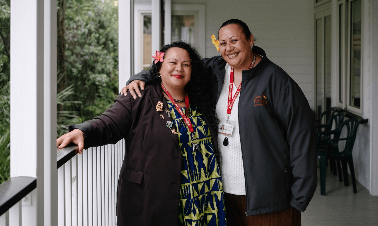 Two women stand side by side on a porch, smiling at the camera. Both wear lanyards and flower hair accessories, with greenery visible in the background. One has her arm around the other's shoulders.