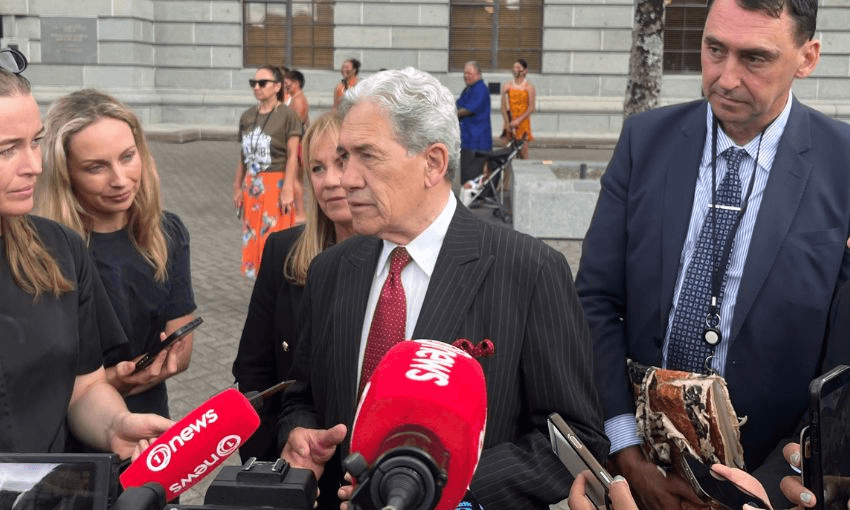 Winston Peters looks to his right while standing in front of a row of microphones, next to MPs Jenny Marcroft and Jamie Arbuckle.