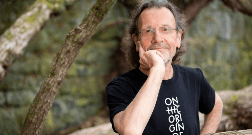 A photograph of Brian Boyd who is a middle aged man with shoulder length hair. He is wearing glasses and a black T-shirt. He is outside, leaning against a tree.