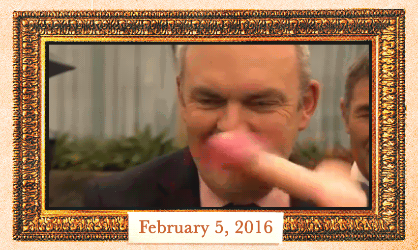 A man in a suit is lightly hit in the face with a rubber penis toy. The scene is framed with an ornate gold border, and "February 5, 2016" is written on a label at the bottom.