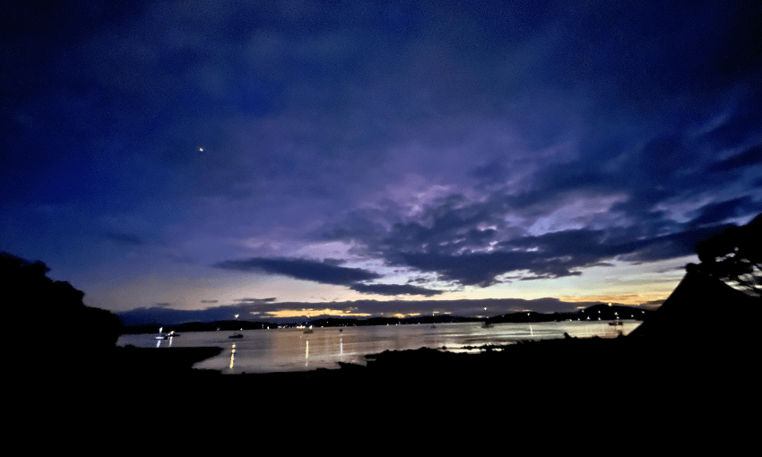 A tranquil morning scene shows a dark shoreline with a calm body of water reflecting the deep blue and purple hues of a cloudy sunset sky. Small lights from distant boats or buildings dot the horizon.