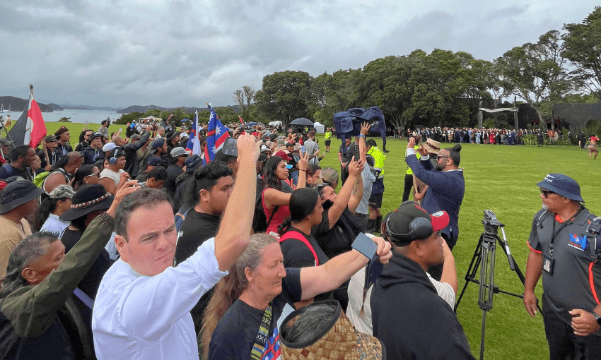 A large crowd stands outdoors on a grassy field, many raising their fists or holding phones. Some people wave flags. Trees and a cloudy sky are in the background. A few security personnel and a camera are visible on the right.