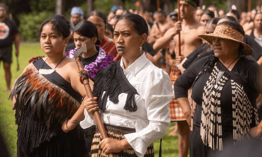 A group of Māori women and men in traditional clothing stand outdoors during a cultural event. One woman in front holds a staff and has traditional facial markings, while others wear feathered cloaks and woven patterns.
