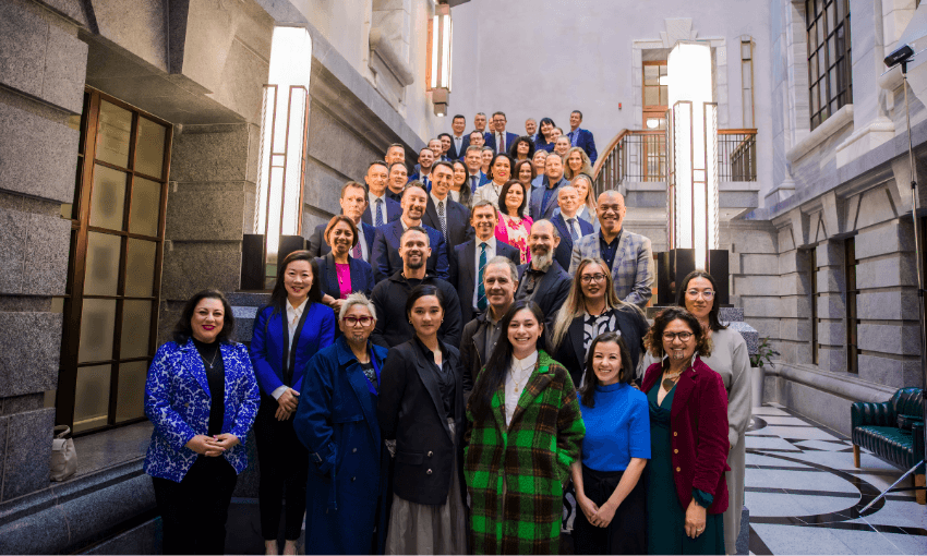 A large group of professionally dressed people stand on a grand staircase in a bright, elegant building, smiling at the camera. The group includes men and women of diverse backgrounds.