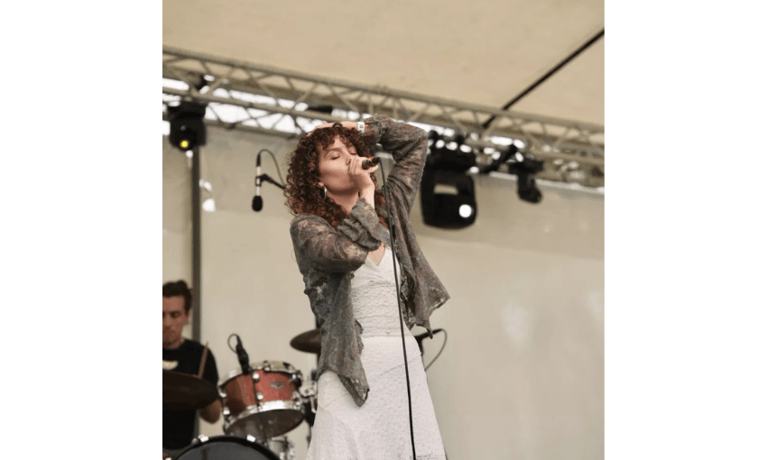 A photo of a young woman wearing a light dress and grey cardigan singing into a microphone on a stage.