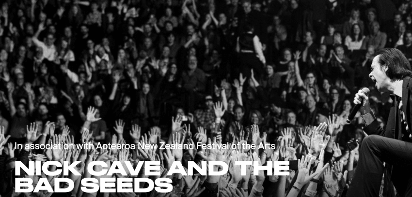 A black and white photo of a large and dense crowd at a concert, with the musician Nick Cave singing into a microphone.