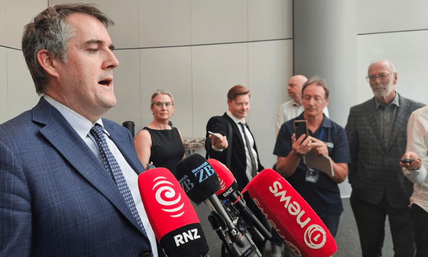 A man in a suit speaks at a press conference while several microphones and reporters with recording devices and notepads are positioned in front of him.
