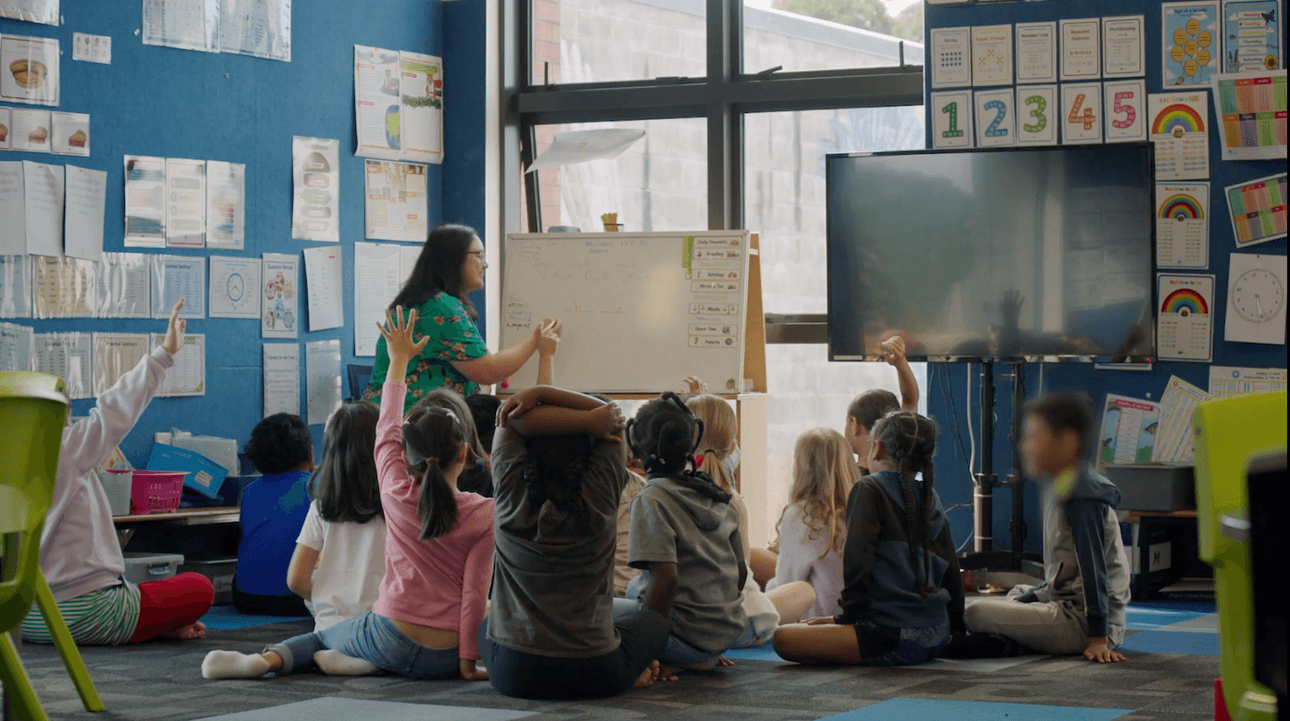 A teacher stands at a whiteboard in a classroom, engaging a group of seated children, some of whom are raising their hands. Colorful posters and a large screen are visible on the blue walls.
