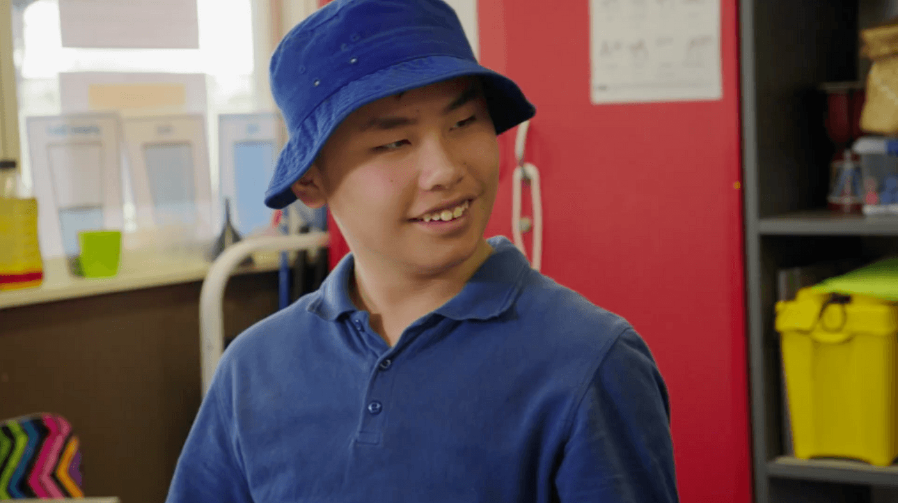 A young person wearing a blue bucket hat and blue shirt smiles while looking to the side in a colorful classroom setting.