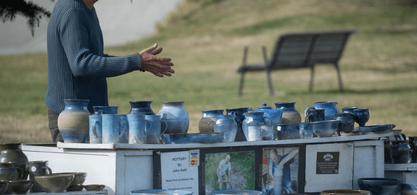 A photo of an outdoor table laden with ceramics. A person is standing behind the table, gesturing.