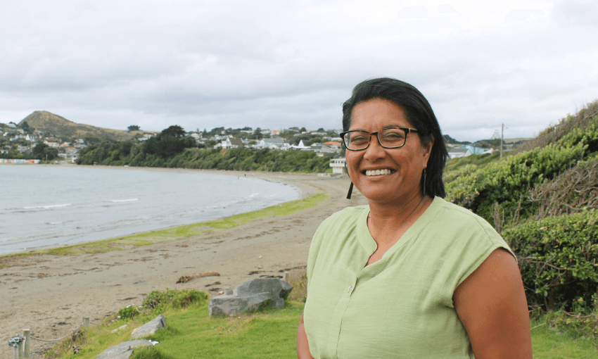 A woman with brown skin and black hair, wearing glasses, smiles at the camera. Behind her is a sandy beach and coastline with clouds above