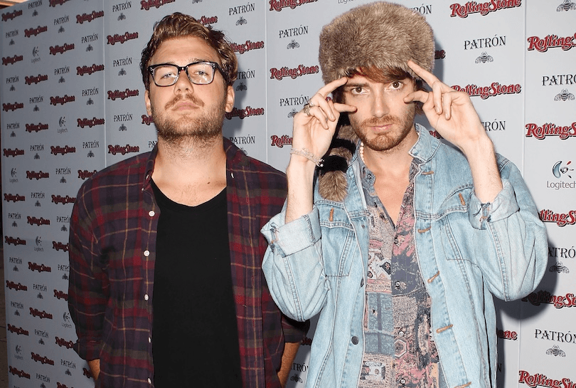Two men pose in front of a Rolling Stone and Patrón step-and-repeat backdrop; one wears glasses and a black shirt, the other wears a fur hat, patterned shirt, and denim jacket, holding his hands up near his face.