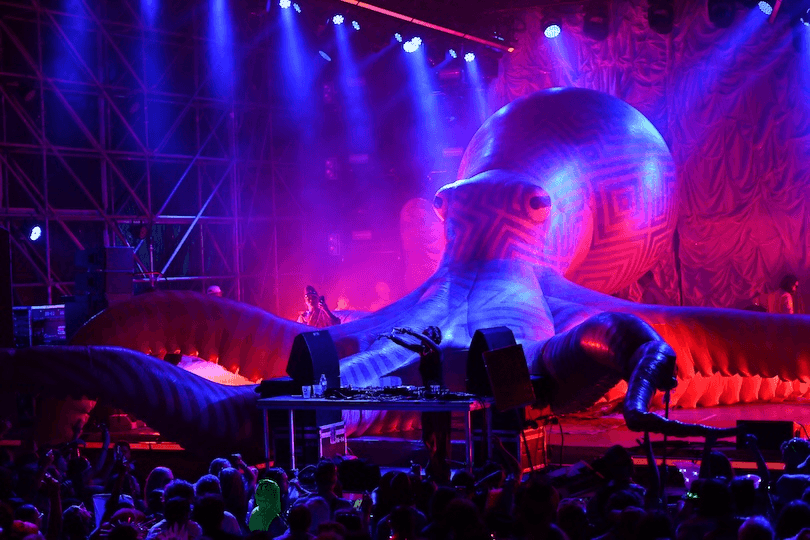 A DJ performs on stage in front of a large, colorful inflatable octopus at a concert, with bright stage lights illuminating the crowd and creating a vibrant atmosphere.
