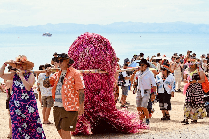 A group of people in colorful, festive outfits carry a large, sparkly pink furry structure on poles along a beach, with the sea and mountains in the background and a crowd watching.