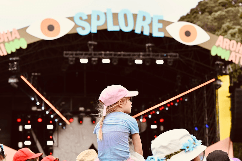 A young girl in a pink cap sits on someone's shoulders at an outdoor music festival, facing a large stage with a colorful