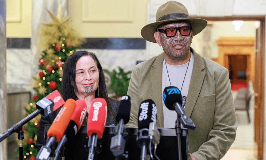 Two people with traditional Māori chin tattoos stand at a podium surrounded by microphones, giving a press conference in an indoor setting with a Christmas tree visible in the background.