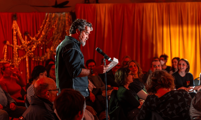 A man stand in front of a microphone and reads from aa card he is holding in his hand. Behind him are the theatre audience