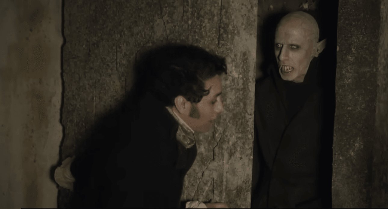 A man in old-fashioned clothing stands near a stone wall, recoiling in fear from a bald, pale figure with sharp teeth and dark sunken eyes emerging from the shadows.