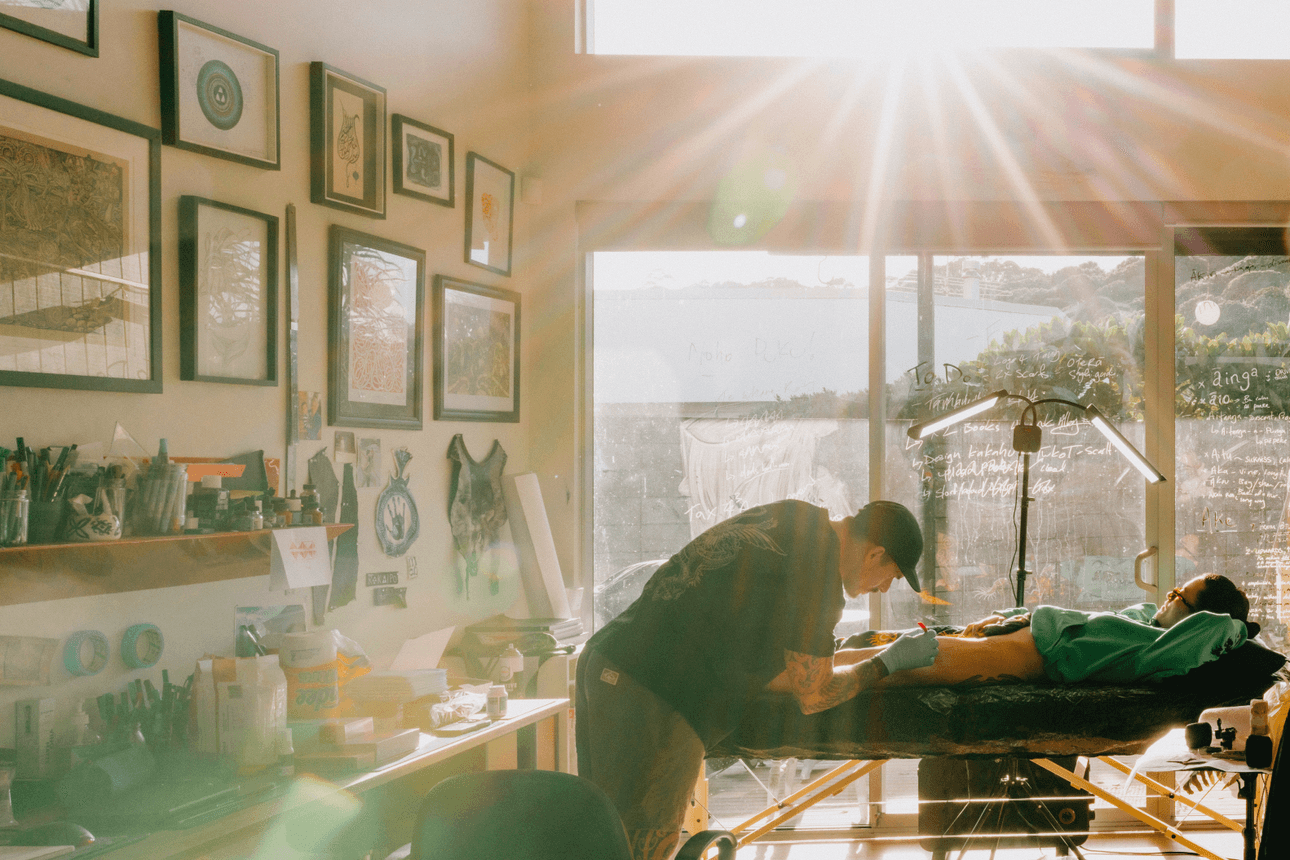 A tattoo artist works on a client lying on a table in a sunlit studio, with rays of light streaming through large windows behind them.