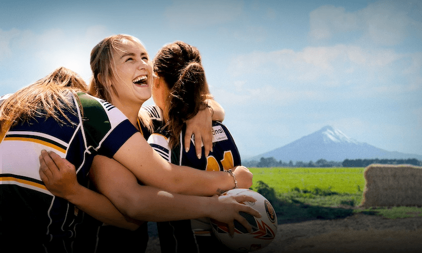 Three women hug on the rugby field with Taranaki Maunga in the background