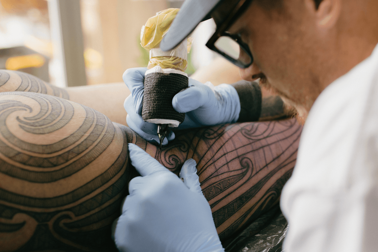 A tattoo artist wearing gloves works on a person's arm, which is covered in intricate, swirling patterns. The image is in black and white, focusing on the tattoo process and detailed body art.