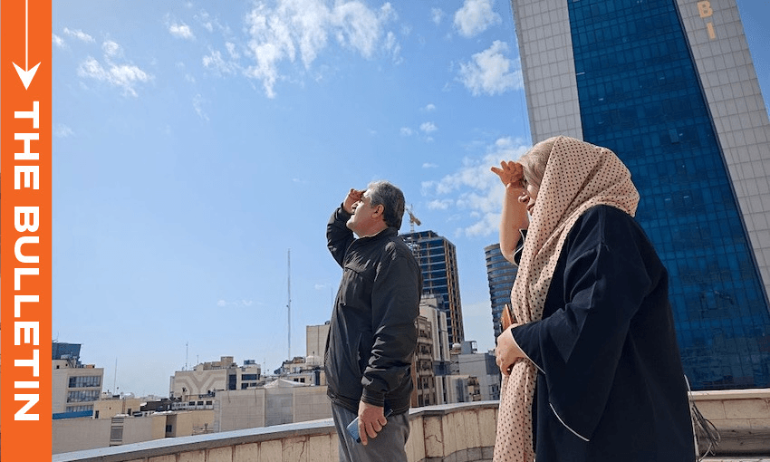 A man and a woman on a rooftop look up at the sky, shielding their eyes with their hands. Tall buildings and a blue sky with clouds are in the background. An orange column reads "THE BULLETIN" on the left side.