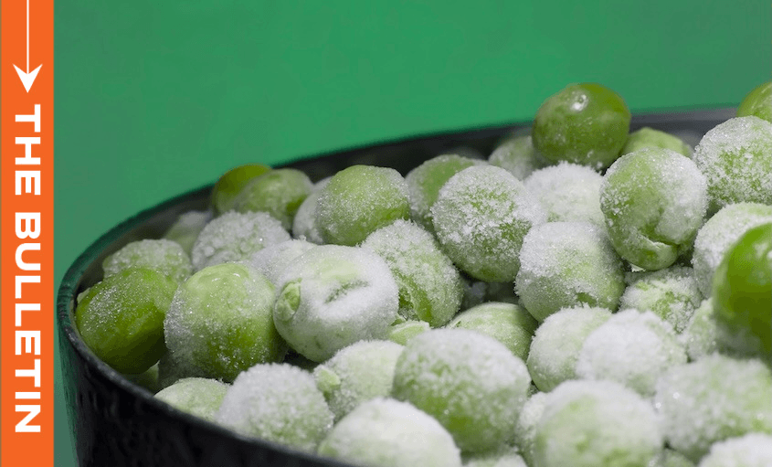 A black bowl filled with frozen green peas, covered in frost, is shown against a green background. An orange vertical banner on the left side reads "THE BULLETIN" in white text.