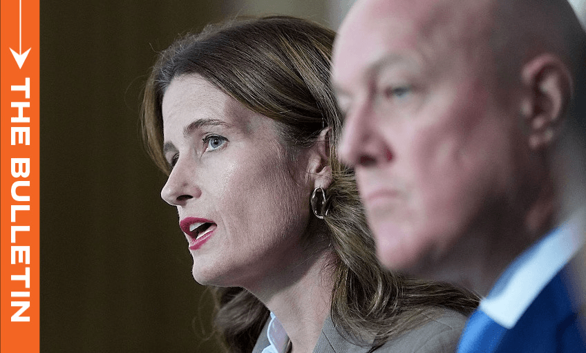 Two people in business attire speak at an event, with a woman in focus on the left and a man out of focus on the right. An orange vertical banner on the left reads "THE BULLETIN.