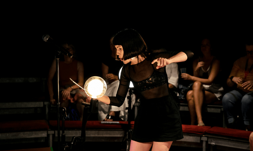 A production photo from a live theatre show that has a woman in a sleek black dress and black bob hair cut gesturing on stage in front of a crowd.