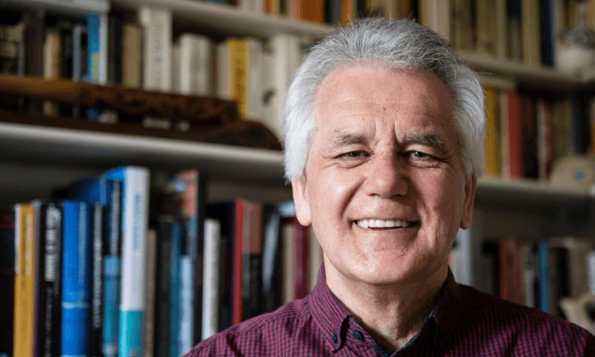 An older man with white hair and a red checkered shirt smiles in front of bookshelves filled with colorful books.