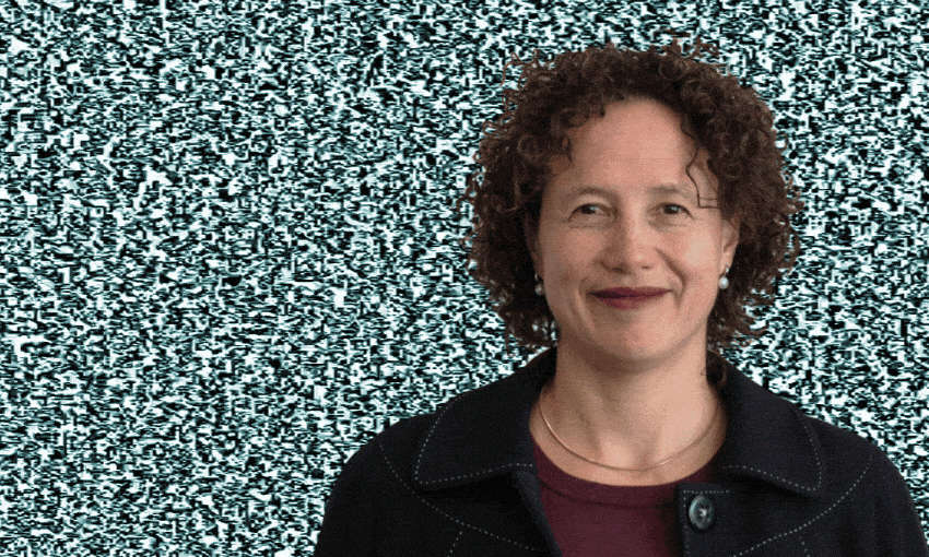 A woman with curly hair and a dark jacket stands smiling in front of a background that looks like black-and-white TV static.